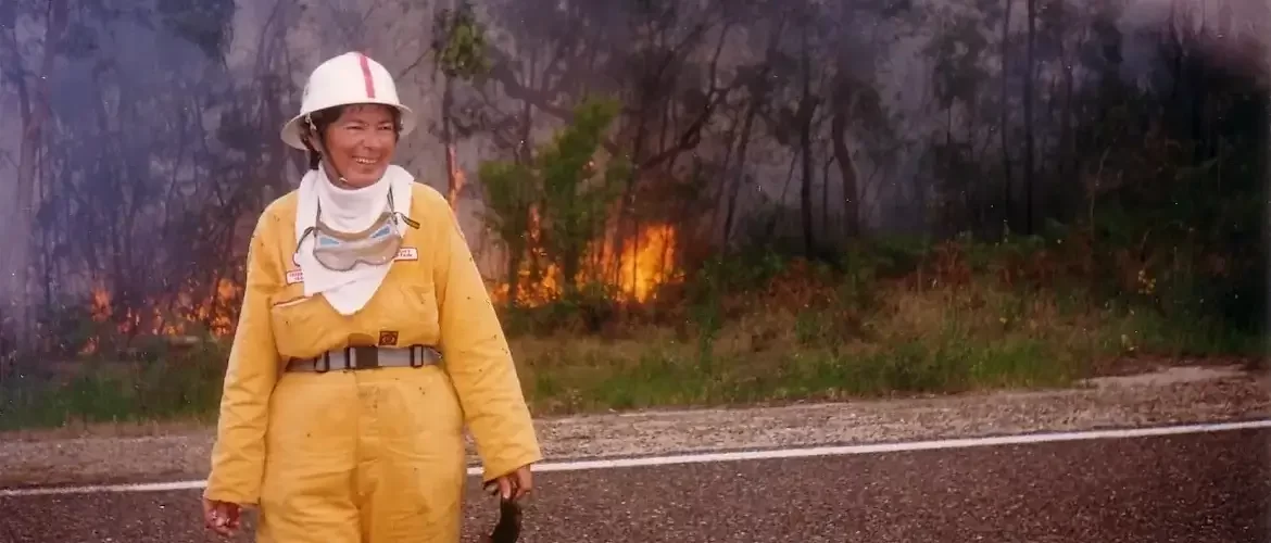 Female firefighter in an orange uniform, with a white hat, in front of a bushfire